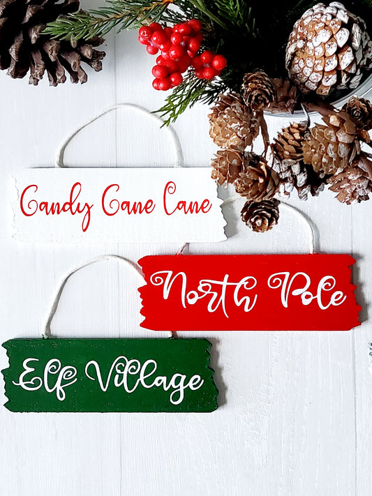 3 wooden hanging décor signs painted each in white, red and green are 2-inches in height and 6.25-inches in length.  Displaying the phrases Candy Cane Lane, North Pole and Elf Village in white. Hand-painted by Snuggle Up Co.