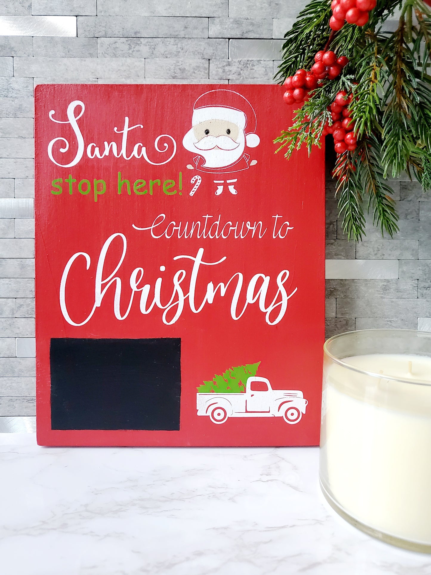 Red wooden sign with white and green lettering that reads Santa stop here! Countdown to Christmas. The sign features a chalkboard area for writing numbers and includes a white truck graphic carrying a green Christmas tree. There is also an illustration of Santa's face with a candy cane in the design.