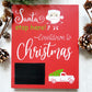 Red wooden sign with white and green lettering that reads Santa stop here! Countdown to Christmas. The sign features a chalkboard area for writing numbers and includes a white truck graphic carrying a green Christmas tree. There is also an illustration of Santa's face with a candy cane in the design.