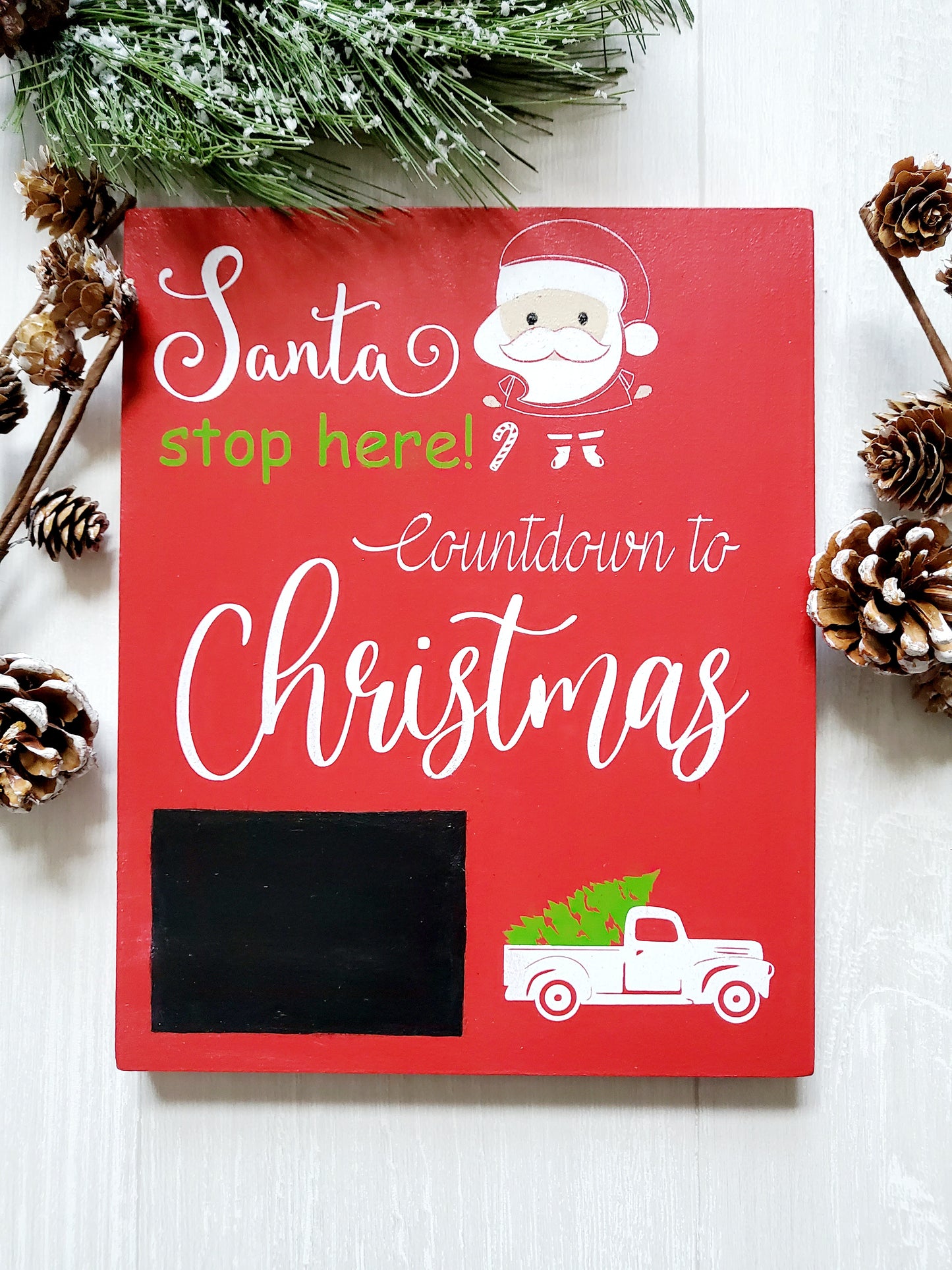 Red wooden sign with white and green lettering that reads Santa stop here! Countdown to Christmas. The sign features a chalkboard area for writing numbers and includes a white truck graphic carrying a green Christmas tree. There is also an illustration of Santa's face with a candy cane in the design.