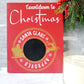 Red wooden sign with white lettering that reads Countdown to Christmas. The sign features a circular gold emblem with the words Santa Claus Approved around a central chalkboard area for writing numbers. The emblem includes silhouettes of Santa in his sleigh being pulled by a reindeer, with snowflakes decorating the design.