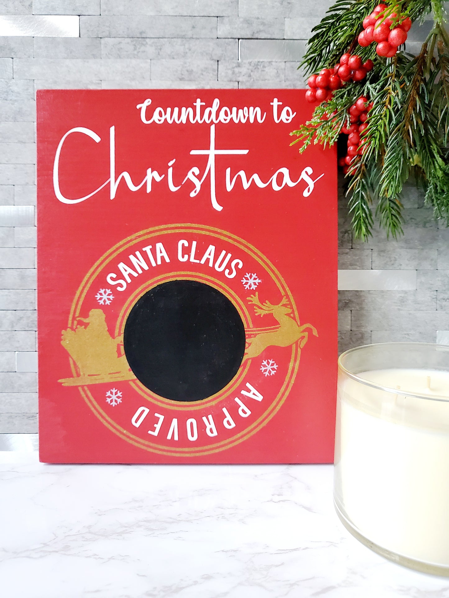 Red wooden sign with white lettering that reads Countdown to Christmas. The sign features a circular gold emblem with the words Santa Claus Approved around a central chalkboard area for writing numbers. The emblem includes silhouettes of Santa in his sleigh being pulled by a reindeer, with snowflakes decorating the design.