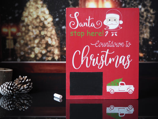 Red wooden sign with white and green lettering that reads Santa stop here! Countdown to Christmas. The sign features a chalkboard area for writing numbers and includes a white truck graphic carrying a green Christmas tree. There is also an illustration of Santa's face with a candy cane in the design.