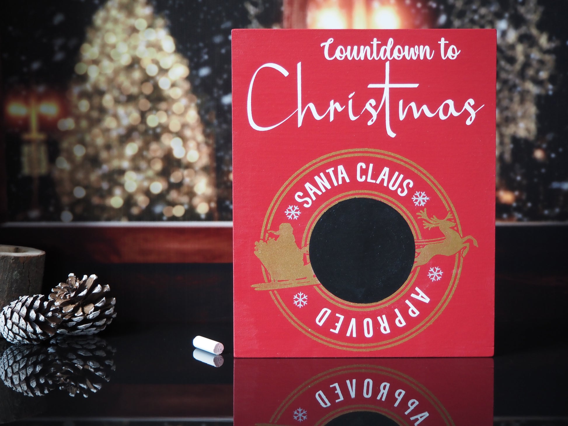 Red wooden sign with white lettering that reads Countdown to Christmas. The sign features a circular gold emblem with the words Santa Claus Approved around a central chalkboard area for writing numbers. The emblem includes silhouettes of Santa in his sleigh being pulled by a reindeer, with snowflakes decorating the design.